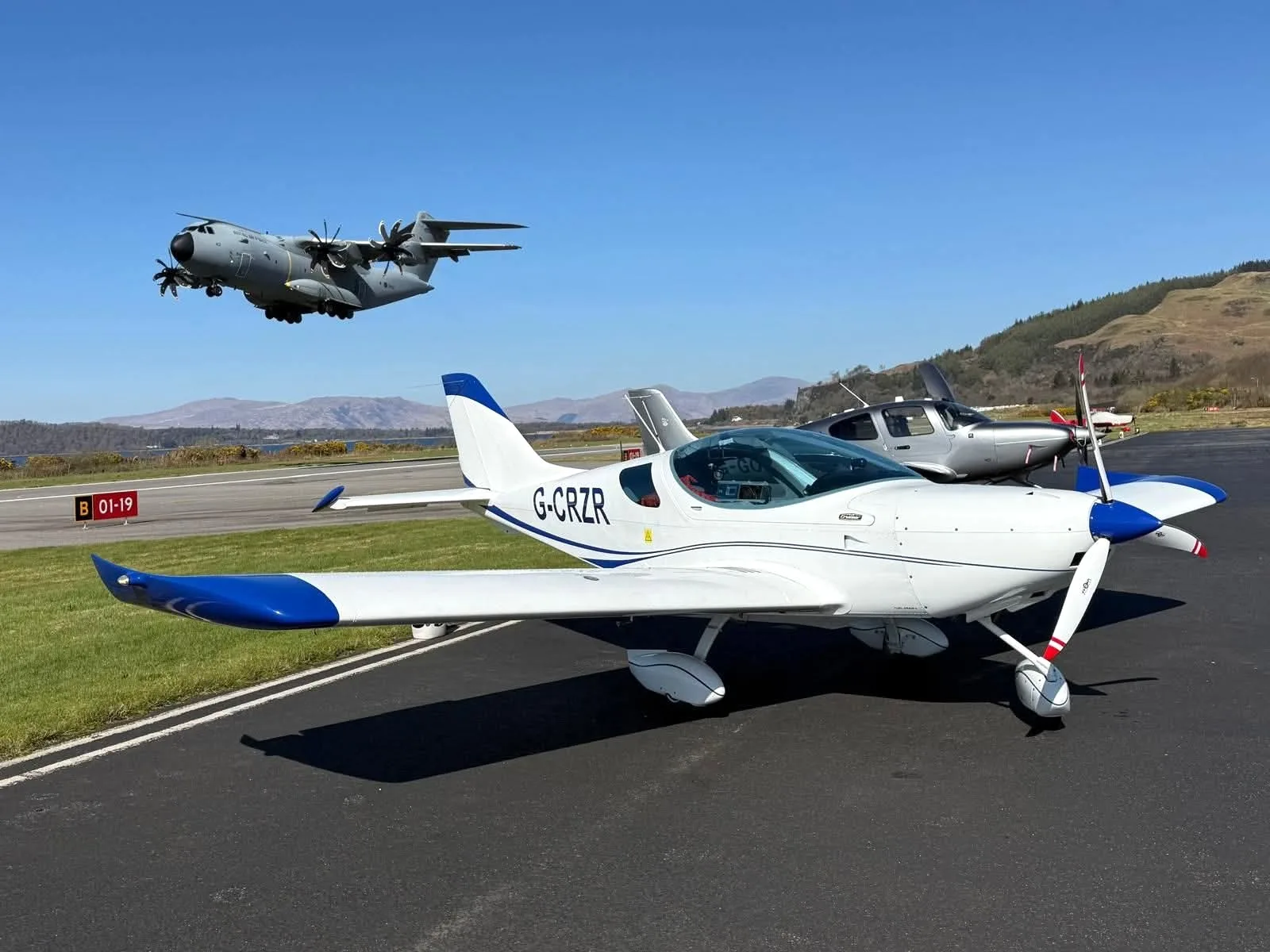 A white and blue light aircraft with registration G-CRZR parked on an airfield, with a large grey military transport plane flying in the background.