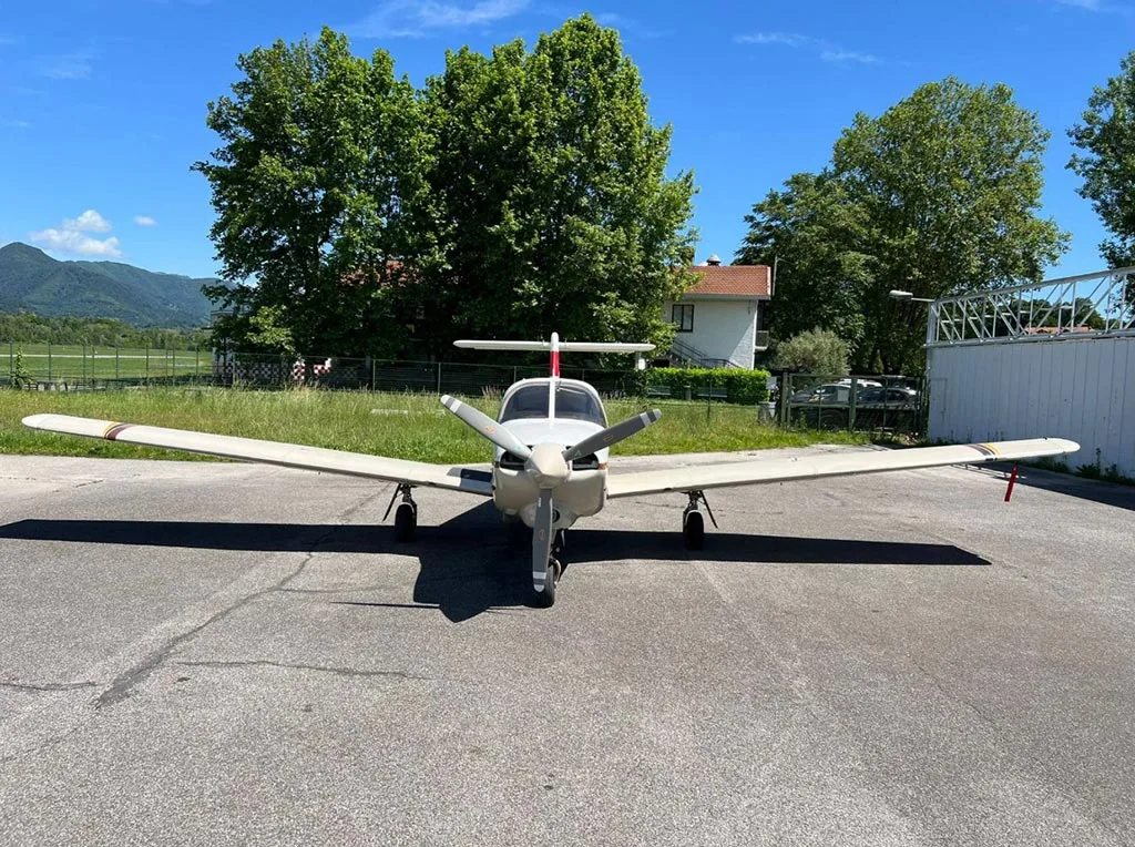 A frontal view of a white and brown Piper Arrow III parked on an asphalt apron with trees and a house in the background.