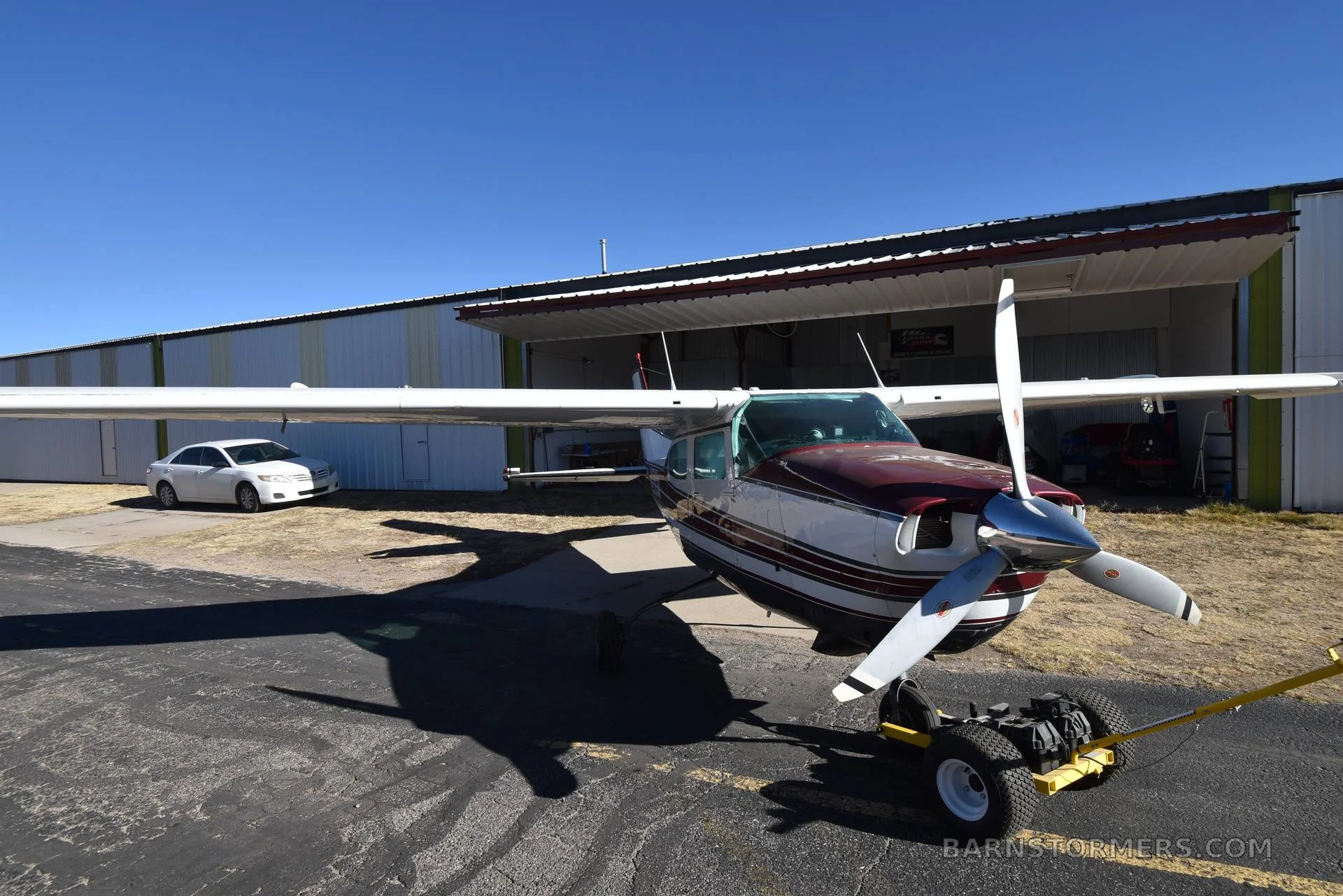 A front view of a white and maroon Cessna 210 aircraft with registration N210W, parked on asphalt with a hangar in the background.
