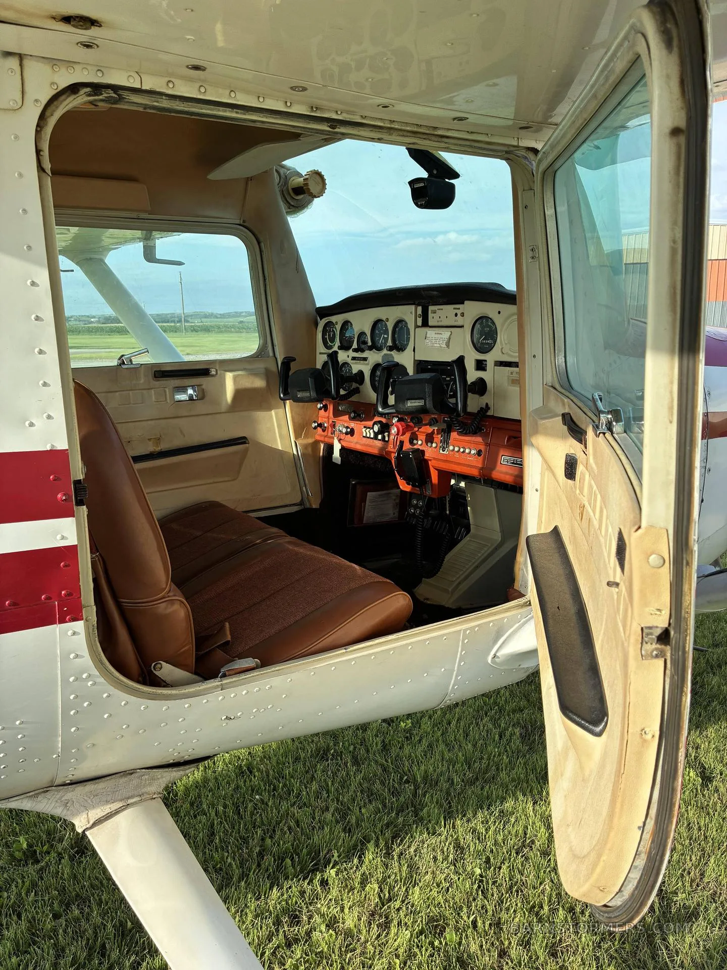 The open door of a Cessna 172 reveals the brown seats and the cockpit from an exterior view.