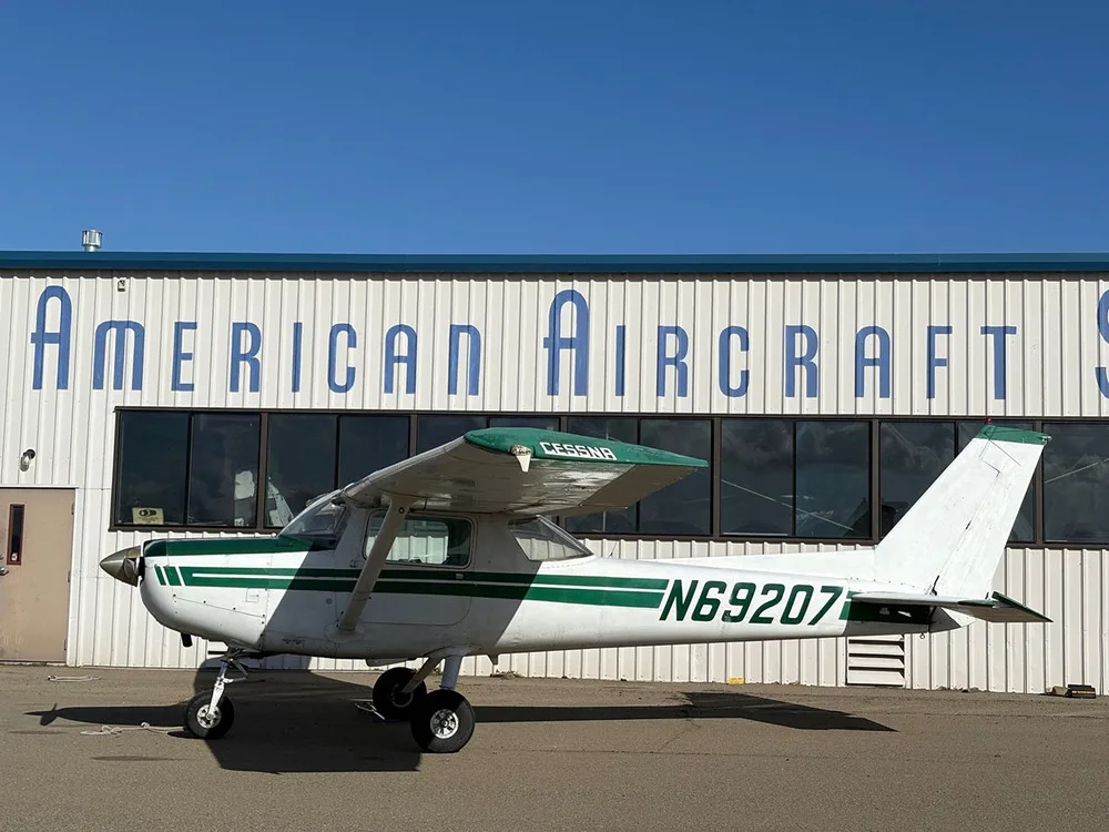 A white and green Cessna N69207 aircraft parked in front of an "AMERICAN AIRCRAFT S" building.