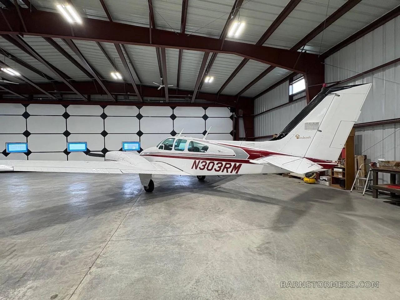 A white twin-engine propeller aircraft with red and grey stripes and tail number N303RM, viewed from the side in a hangar.