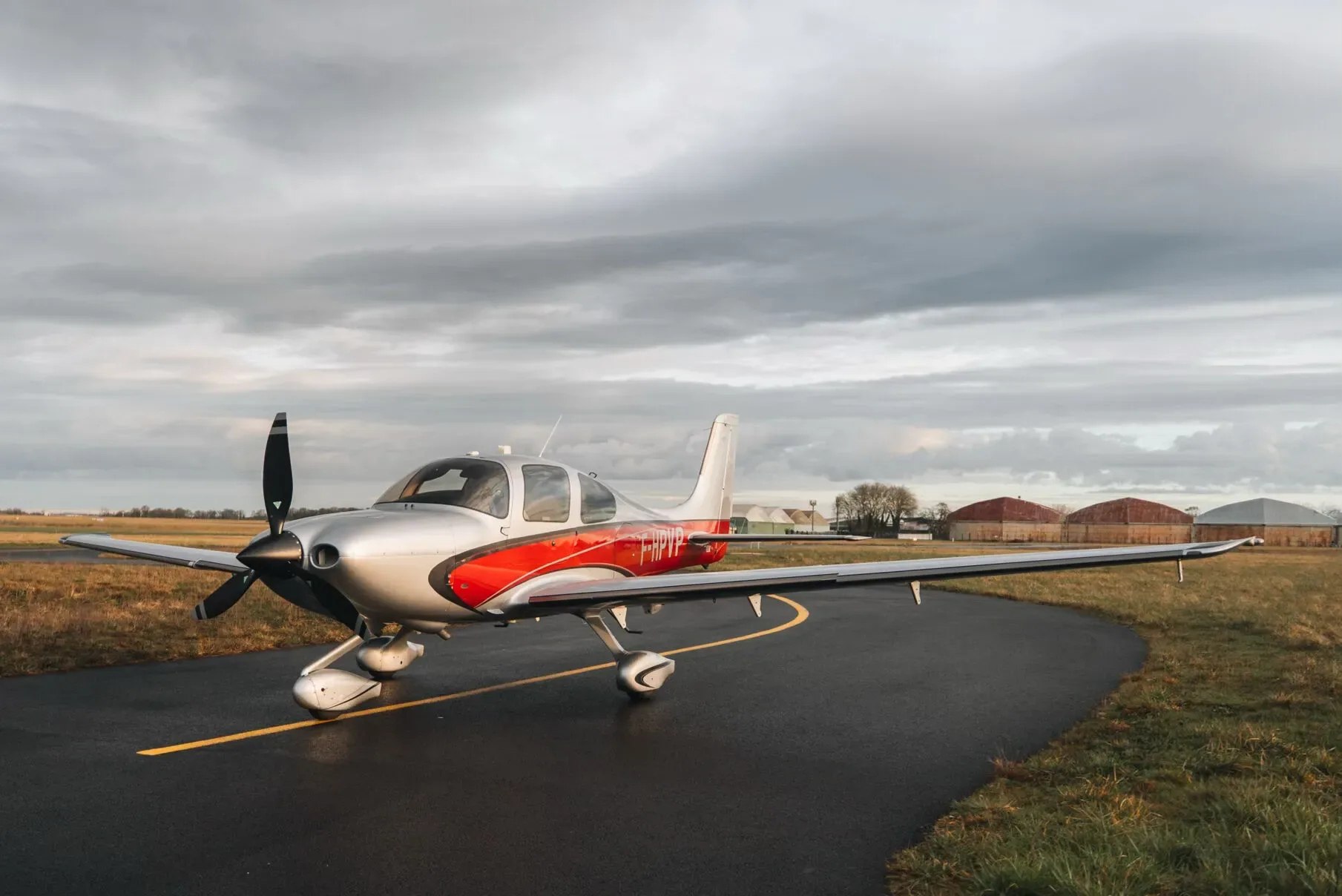 A silver and red Cirrus SR22 aircraft parked on an airfield with a cloudy sky overhead.