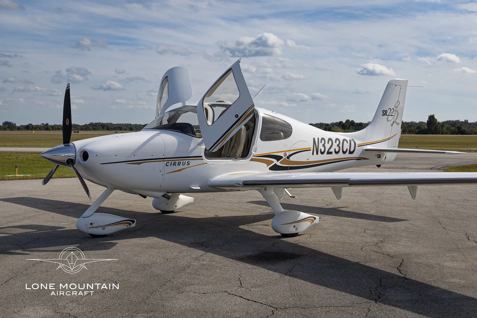 A white Cirrus SR22 aircraft with gold and black accents, registration N323CD, parked on an airfield with its doors open.