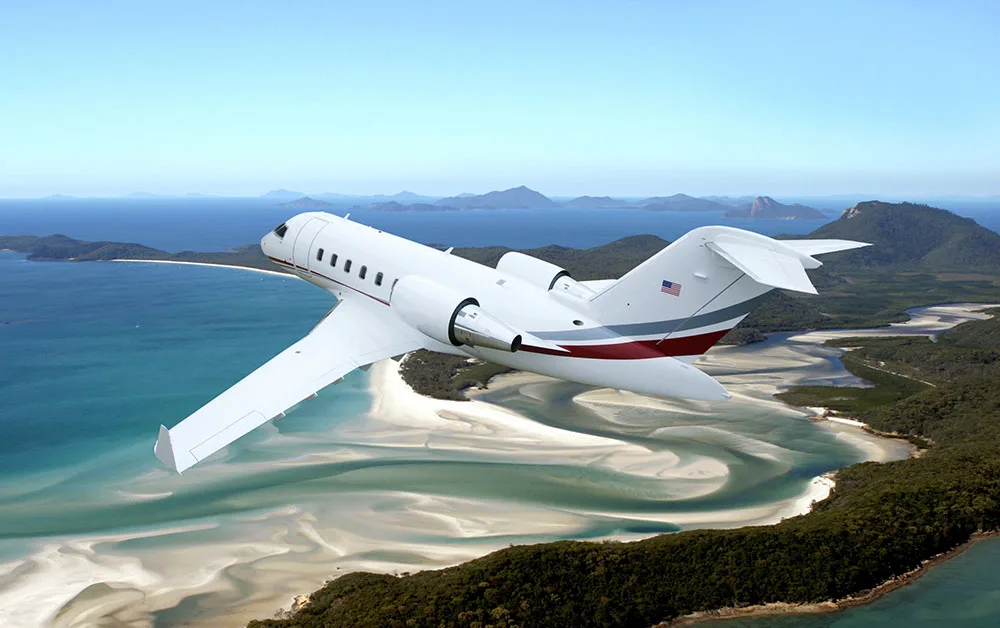 A white business jet with red and grey stripes flies over a tropical coastline with white sand beaches and clear blue water.