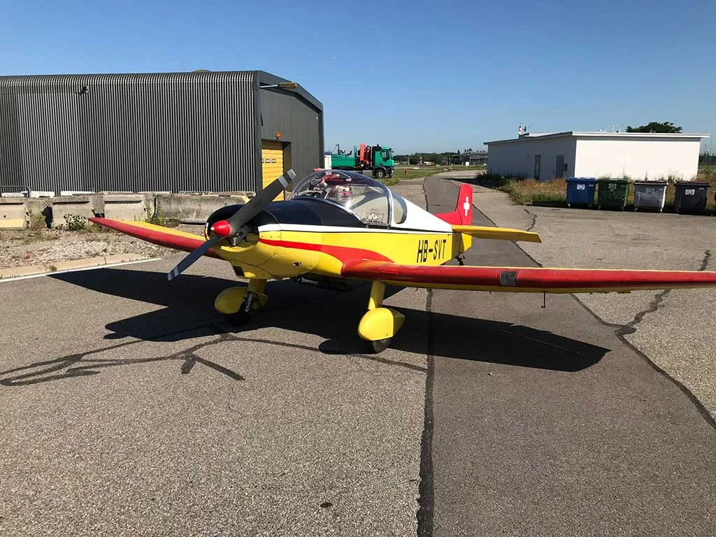 A yellow, red, and black single-engine aircraft (HB-SVT) parked on an asphalt tarmac with buildings and bins in the background.