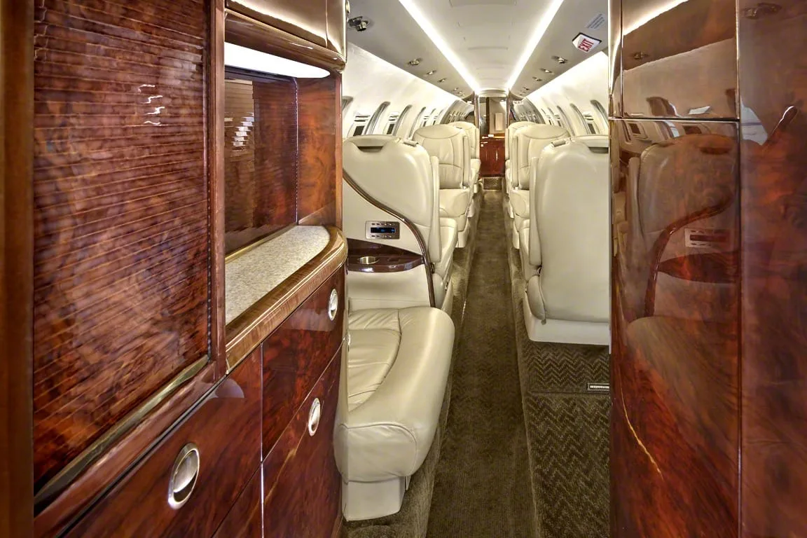 The long interior cabin of a private jet with beige leather seats, dark wood paneling, and overhead lighting.