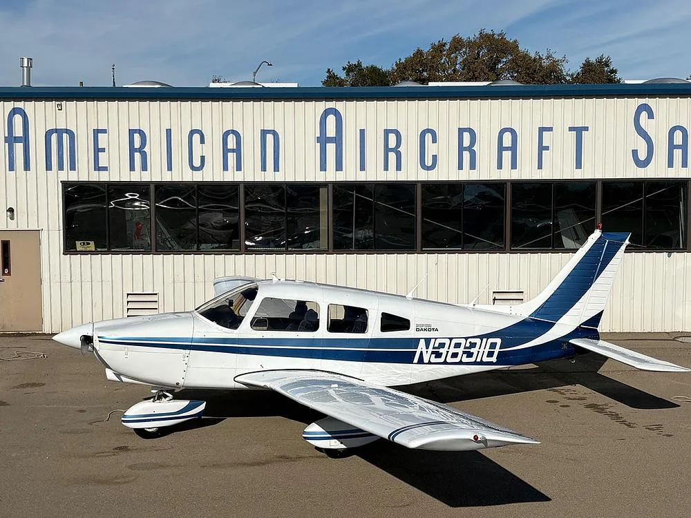 A white Piper Dakota aircraft with blue stripes, registration N3831Q, parked in front of a building labeled "American Aircraft Sales".