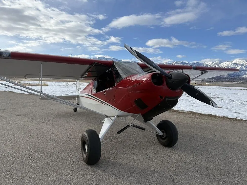 A red and white bush plane with large tires parked on an asphalt runway with snow-capped mountains under a partly cloudy sky.