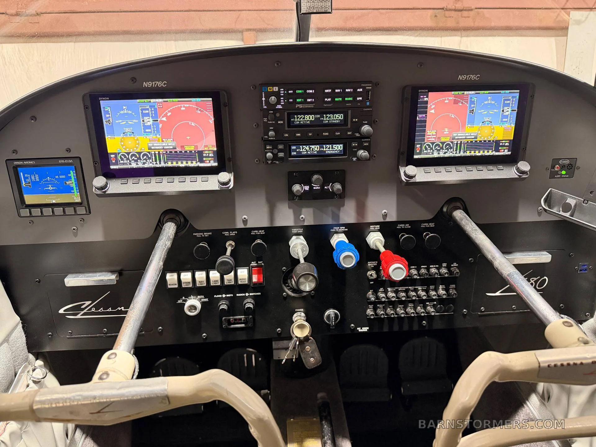 The cockpit of a Cessna 180 aircraft, showing the instrument panel with two large digital displays, various gauges, switches, and control yokes.