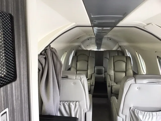The interior cabin of a business jet features multiple rows of grey leather seats and overhead lighting.