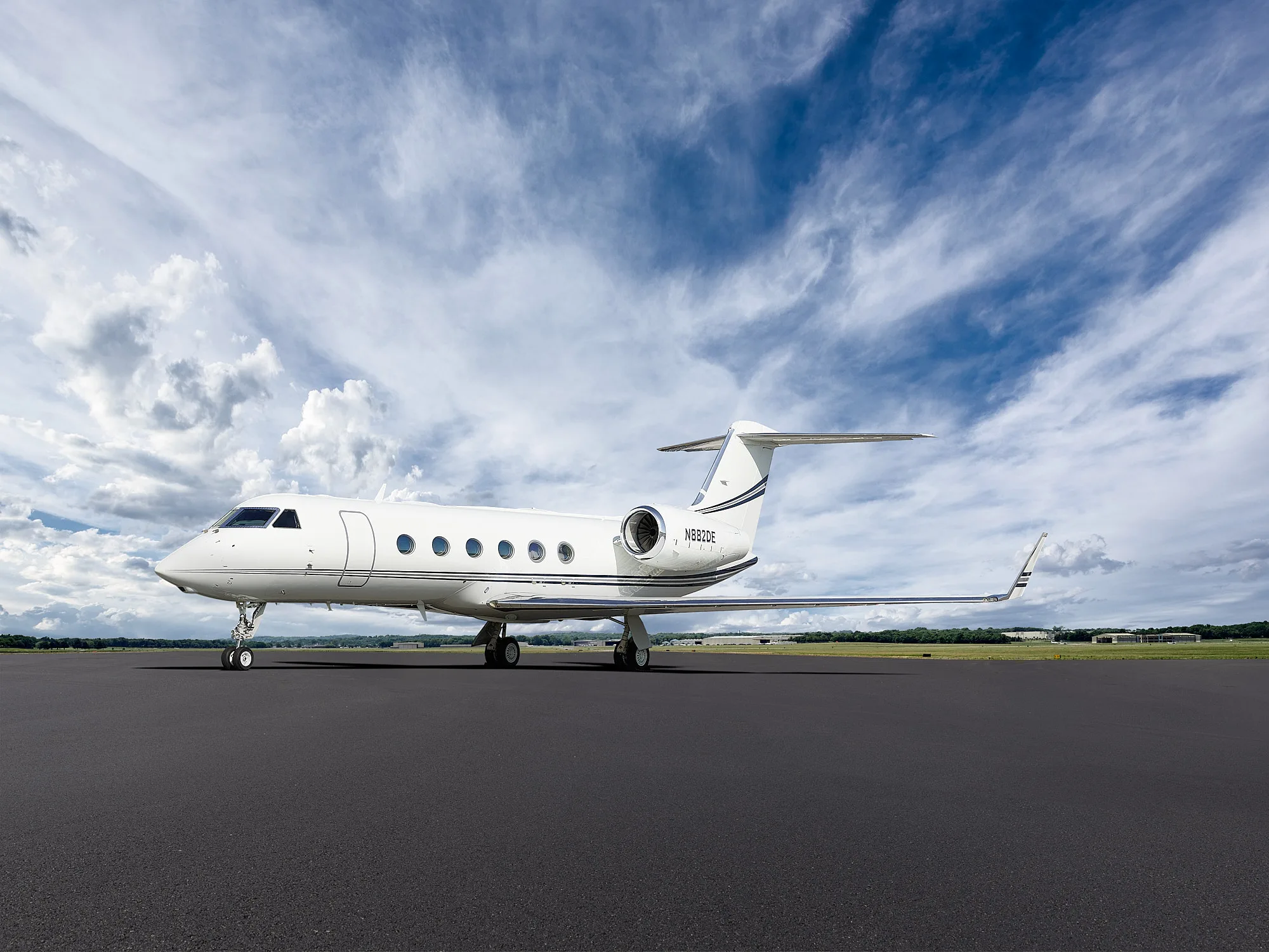 A white Gulfstream business jet with black and grey stripes on the tail is parked on a dark tarmac under a partly cloudy blue sky.