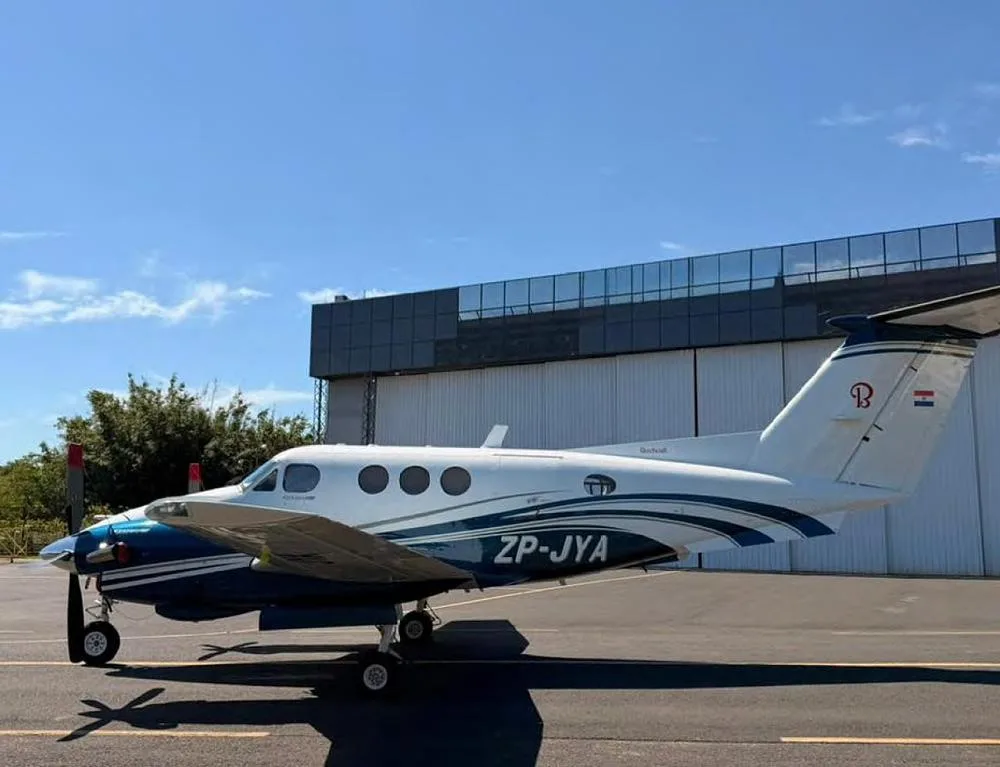A Beechcraft King Air with blue and white livery, registration ZP-JYA, parked on a tarmac next to a modern hangar.