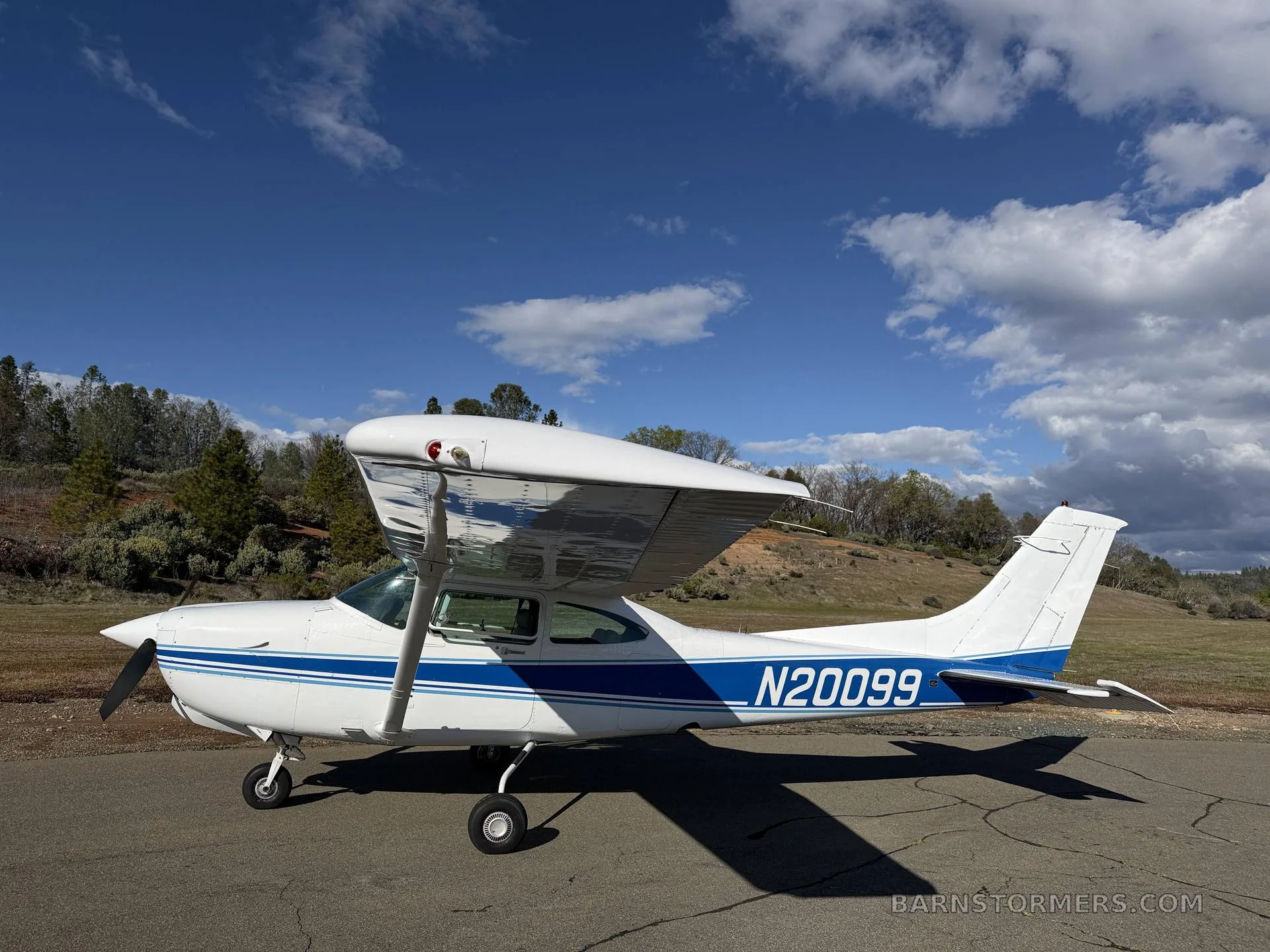 A white Cessna 182 with blue stripes, registration N20099, parked on an asphalt ramp under a partly cloudy sky.