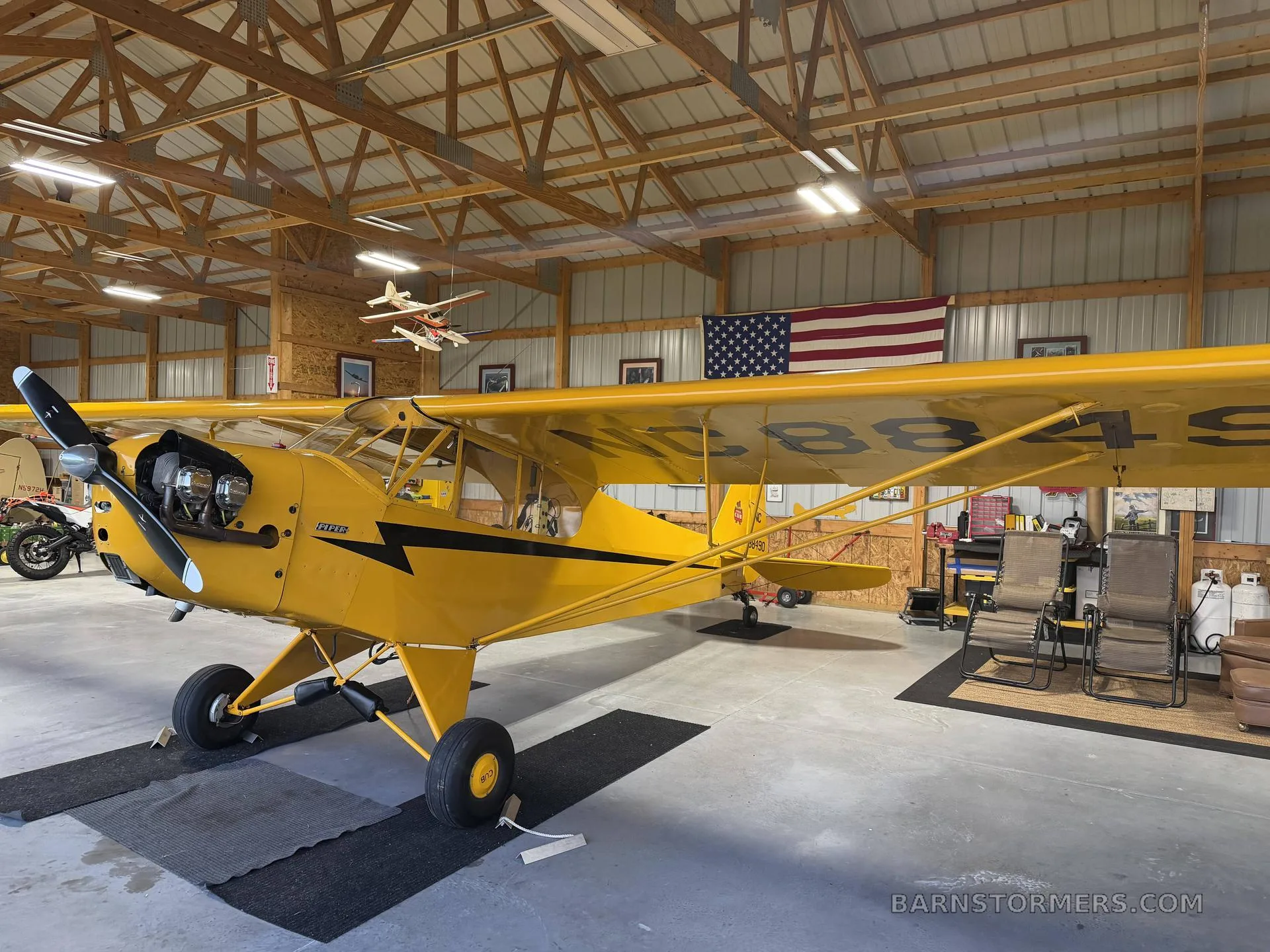 A yellow Piper Cub aircraft with black stripes is parked inside a hangar with an American flag hanging in the background.