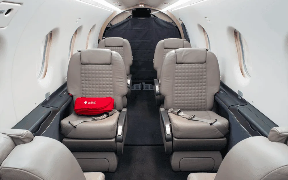 The aircraft cabin features light grey leather seats in a club configuration with a red first aid kit on one seat.