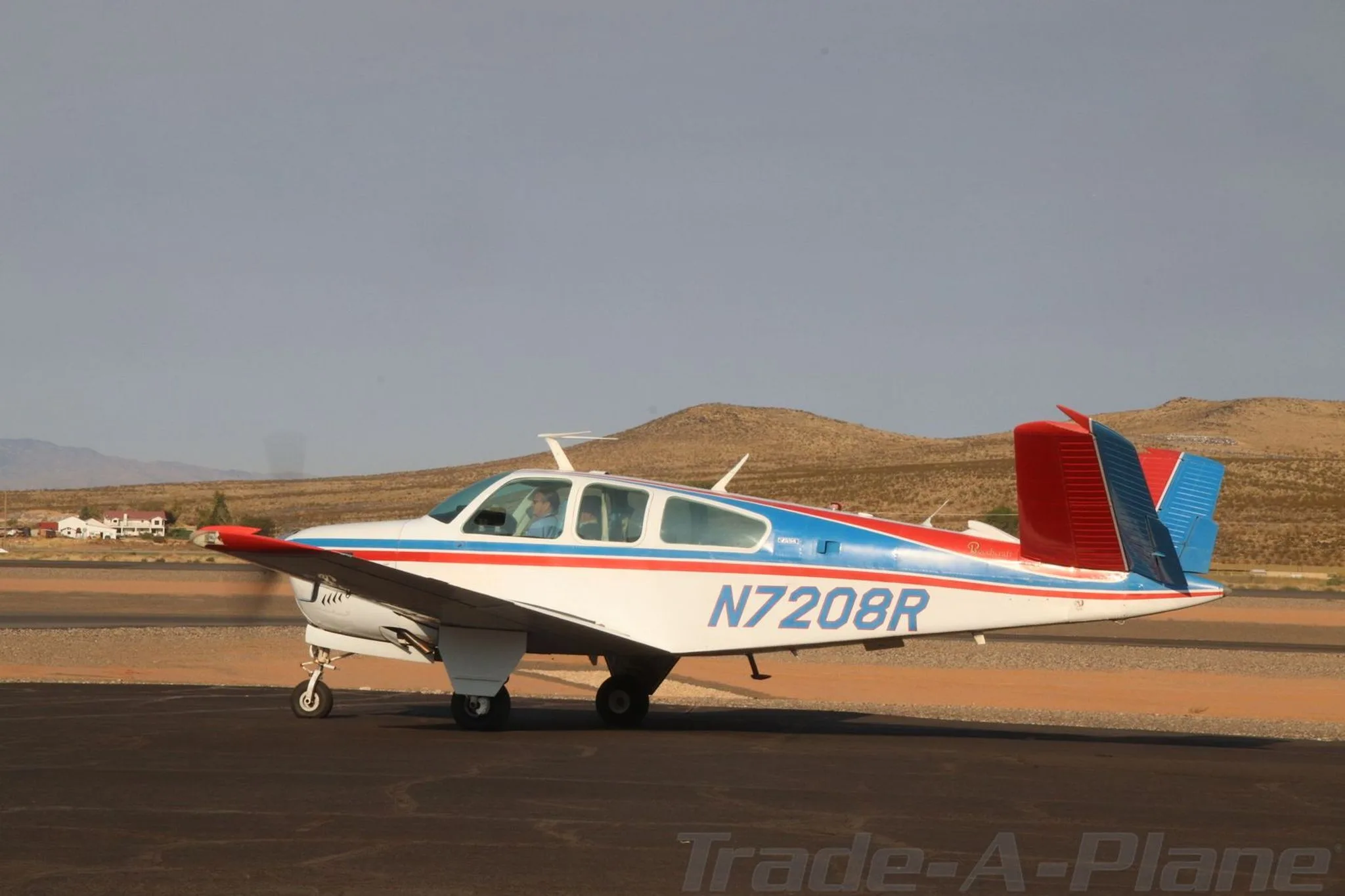 A white, red, and blue Beechcraft Bonanza N7208R is seen from the side on a tarmac with mountains in the background.