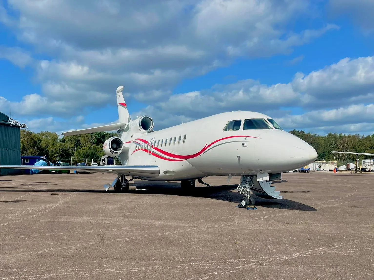 A white Dassault Falcon business jet with red and grey stripes is parked on an airfield under a partly cloudy sky.