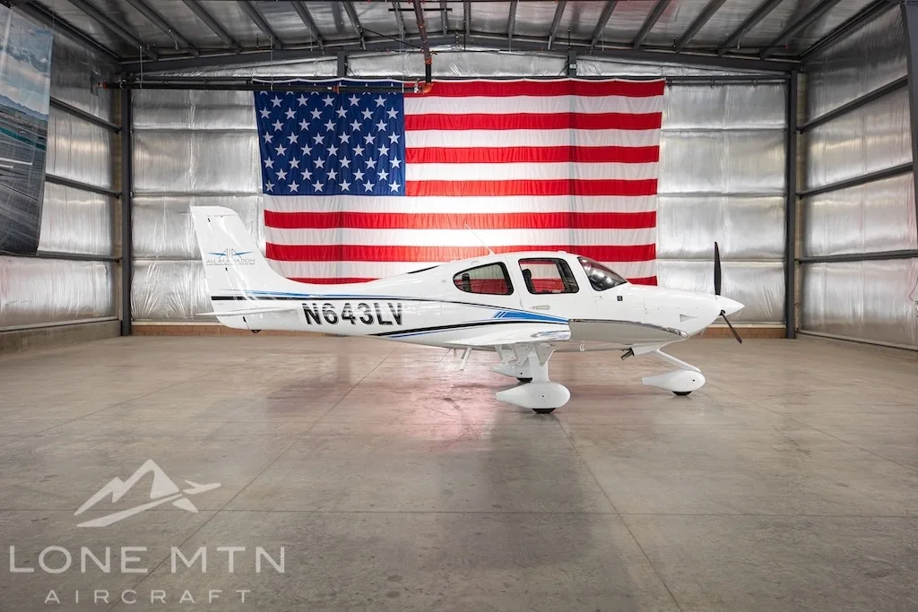 A white Cirrus SR22 aircraft with blue and black stripes, tail number N643LV, parked in a hangar with an American flag in the background.