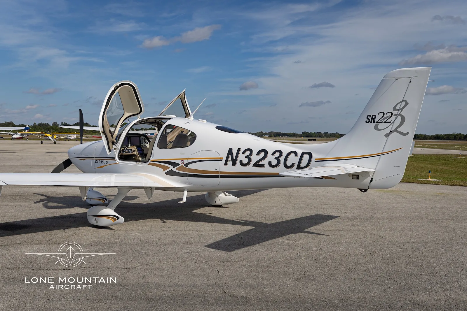 A white Cirrus SR22 aircraft with gold and black accents, registration N323CD, parked on an airfield with its doors open, viewed from the right side.