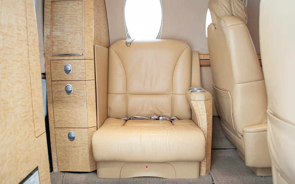 A close-up view of a single beige leather seat and light wood cabinetry inside an aircraft cabin.