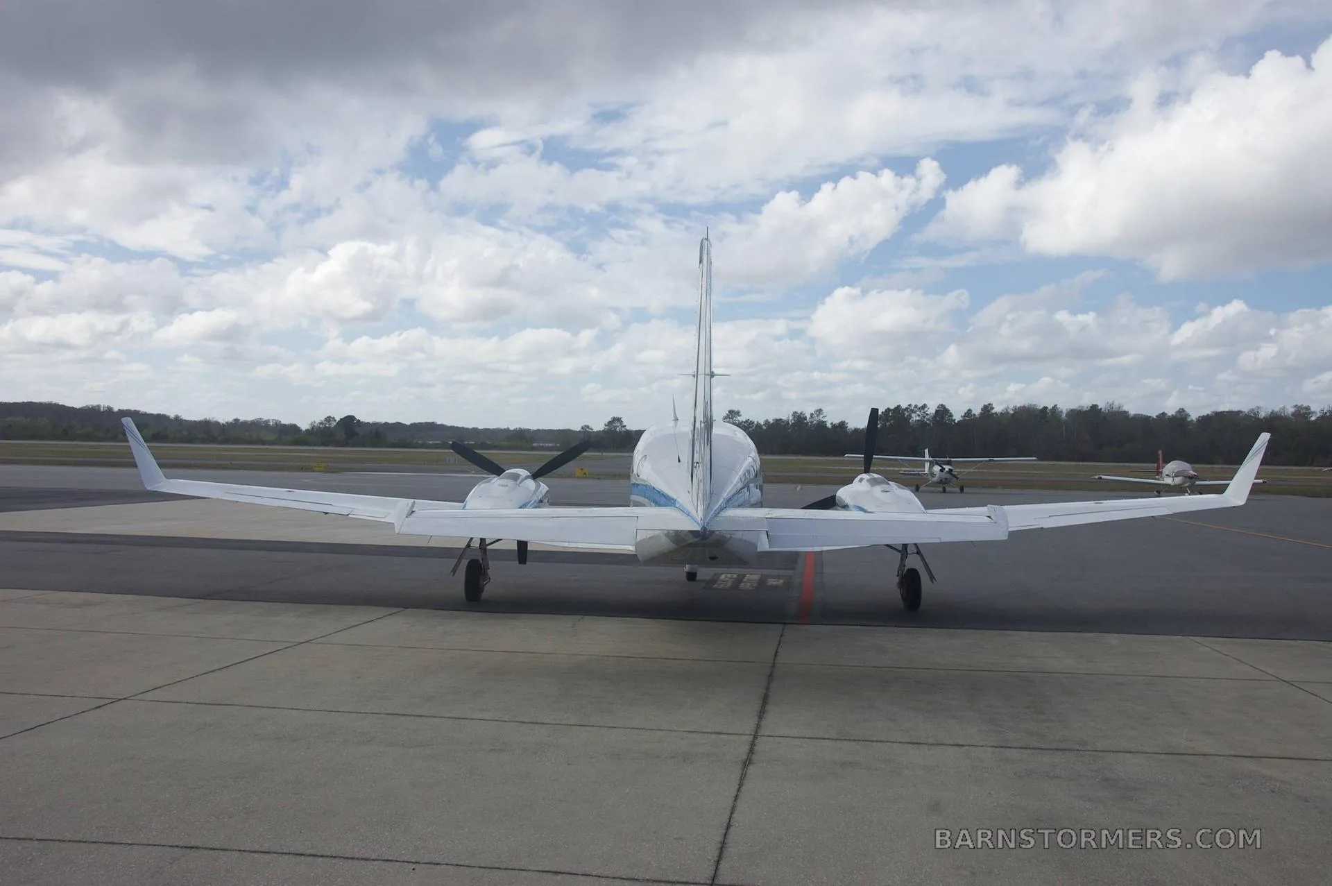 A rear view of a white twin-engine propeller aircraft with blue and grey stripes (N517MH) parked on a tarmac under a cloudy sky.