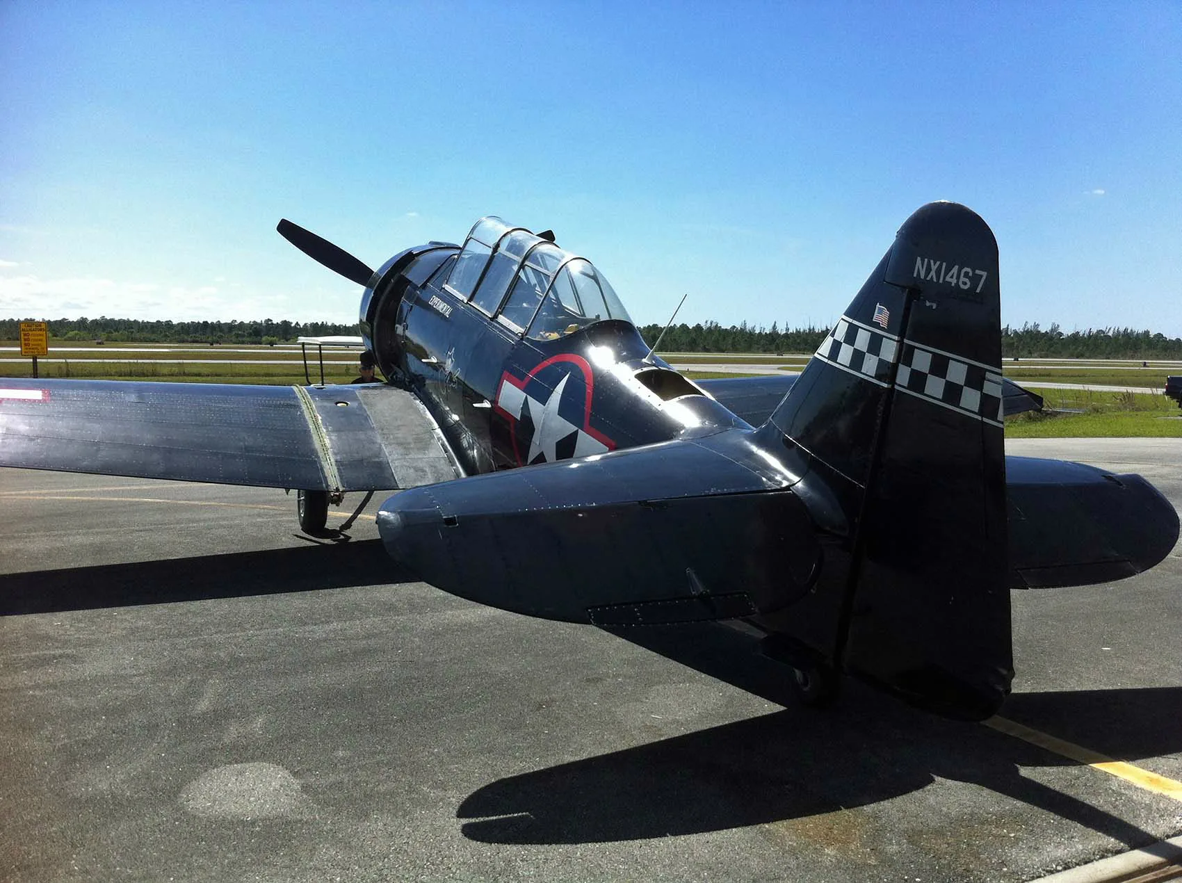 A rear-quarter view of a black warbird aircraft with US star insignia and registration NX1467 on the tail, parked on an airfield.
