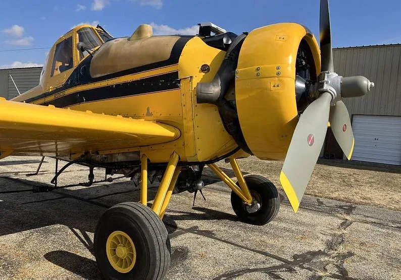 A detailed side view of the front section of a yellow agricultural aircraft, focusing on the engine, propeller, and landing gear.