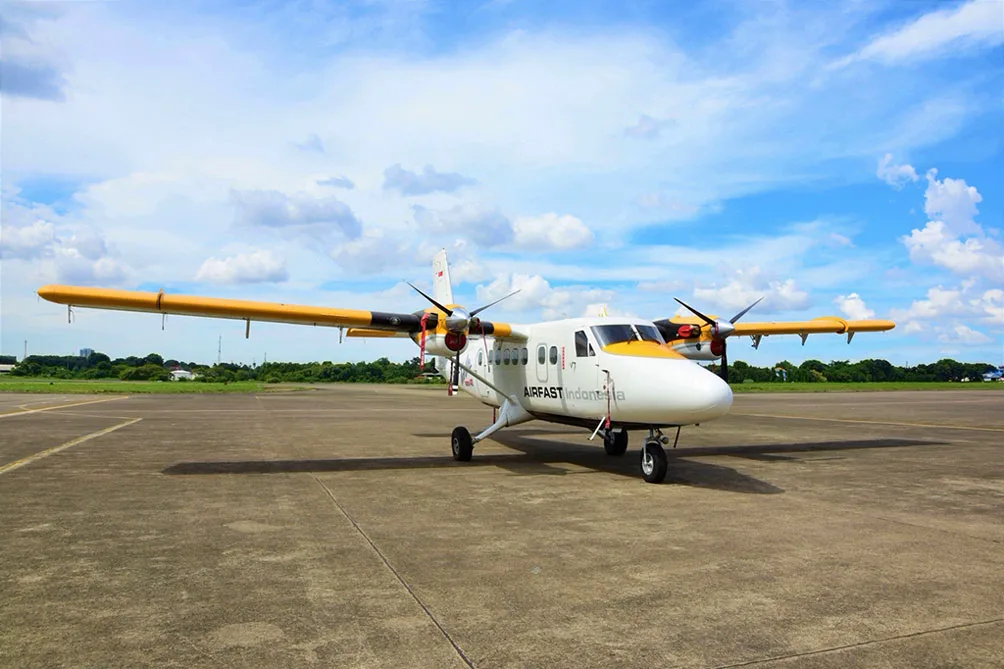 A white and yellow Airfast Indonesia Twin Otter aircraft parked on an airfield under a partly cloudy sky.