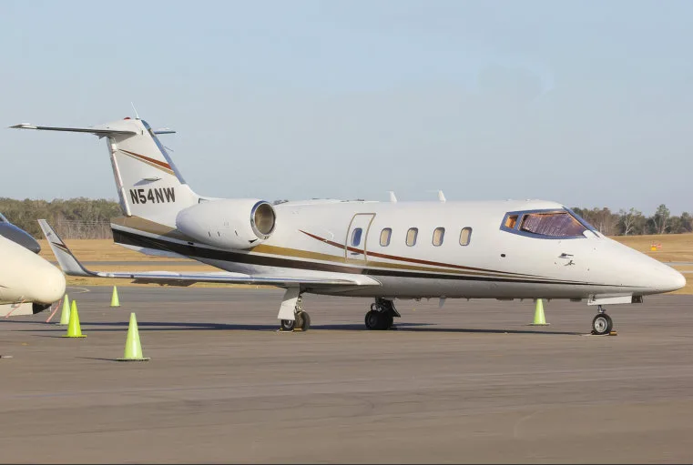 A white Learjet 60 with brown and gold stripes, registration N54NW, parked on an airfield with safety cones.