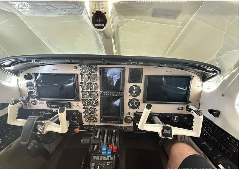 The cockpit of a twin-engine aircraft, showing the instrument panel with analog gauges, two large digital displays, and dual yokes.