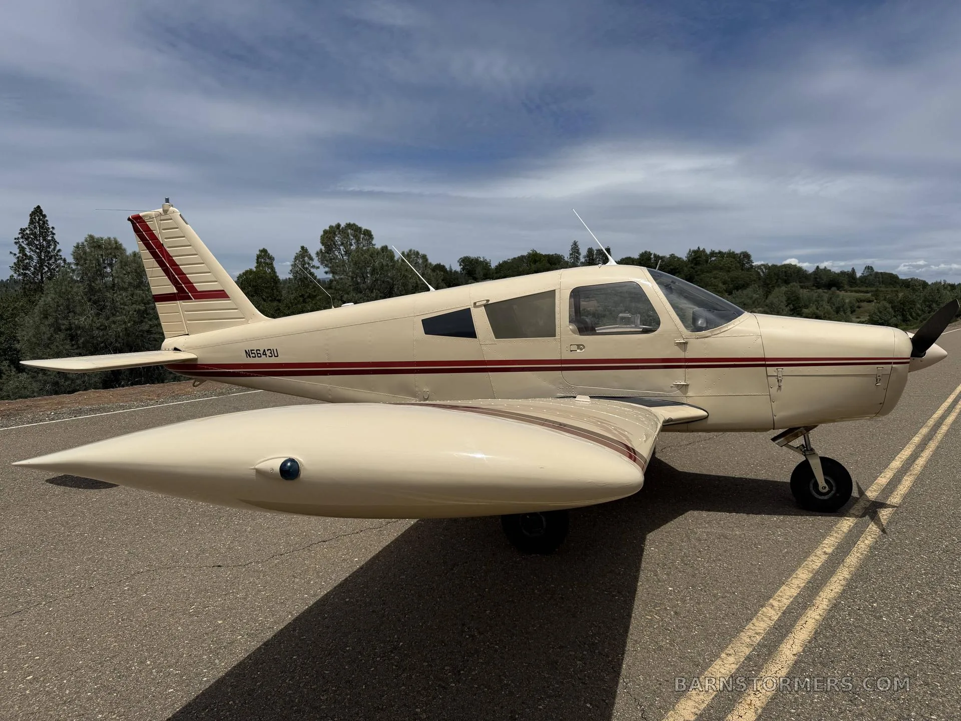 A cream-colored Piper Cherokee with red stripes, registration N5643U, parked on an asphalt surface under a partly cloudy sky, viewed from the front-right side.
