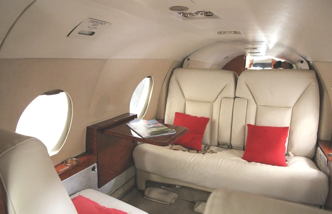 The interior of a private jet cabin featuring cream leather seating, red accent pillows, and a small wooden table.