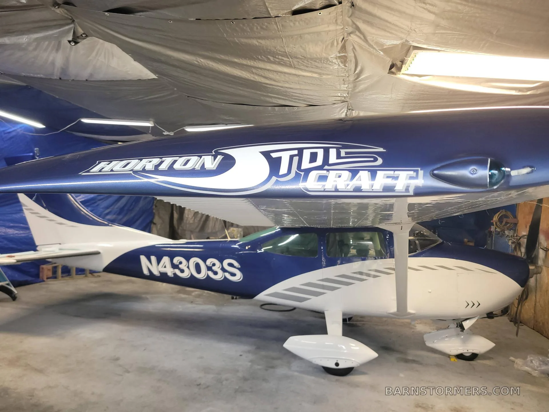 A blue and white Cessna-style aircraft with 'HORTON STOLCRAFT' on the fuselage and tail number N4303S, parked in a hangar.