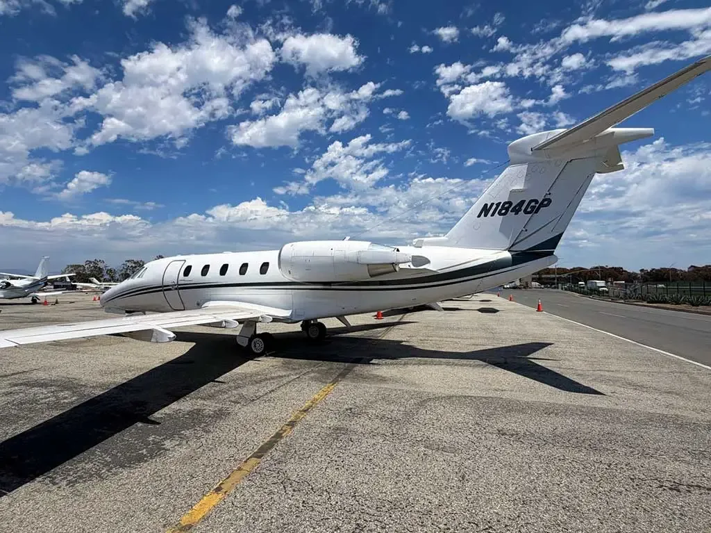 A white Cessna Citation business jet (N184GP) with a dark stripe, viewed from the rear on an asphalt tarmac under a blue sky with scattered clouds.