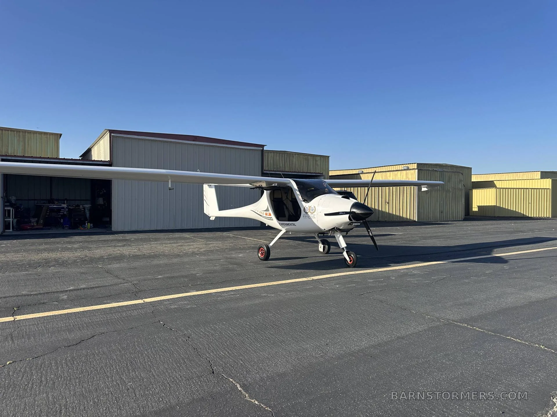 A white light sport aircraft with a black propeller and red wheel hubs, parked on an asphalt tarmac in front of hangars under a clear blue sky.