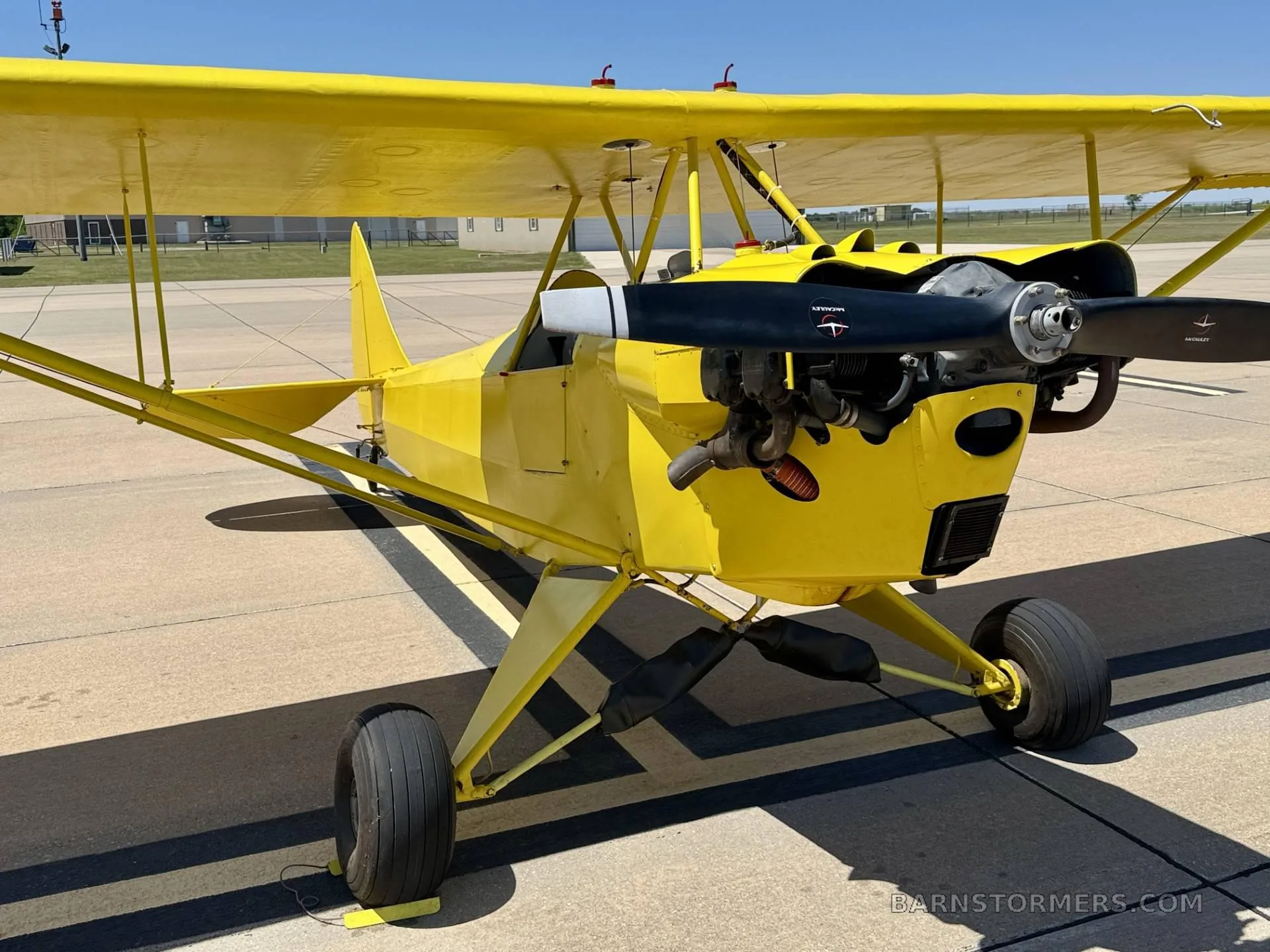 A bright yellow biplane with a visible engine and propeller, parked on an asphalt tarmac under a clear blue sky.