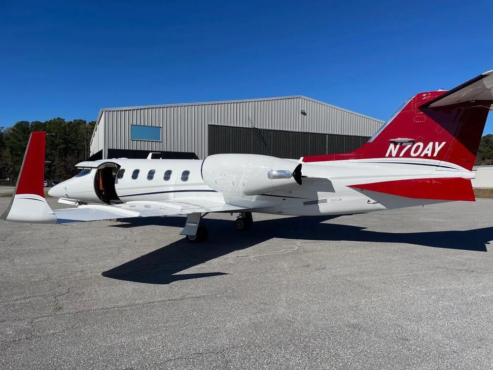 A white Learjet with red tail and accents, registration N70AY, parked with its door open next to a hangar.