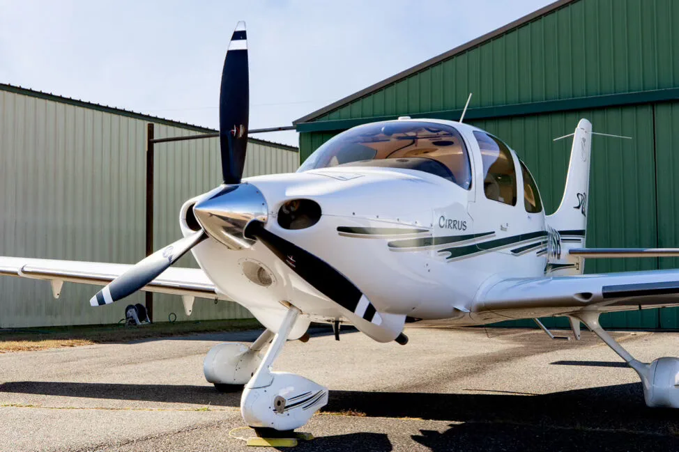 A front three-quarter view of the white Cirrus SR22 aircraft, showing its propeller and green and grey stripe details.