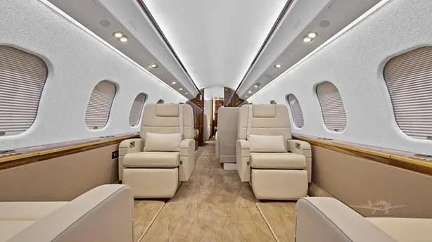 A view down the aisle of a private jet cabin, showing plush cream seats, light-colored walls, and windows with blinds.