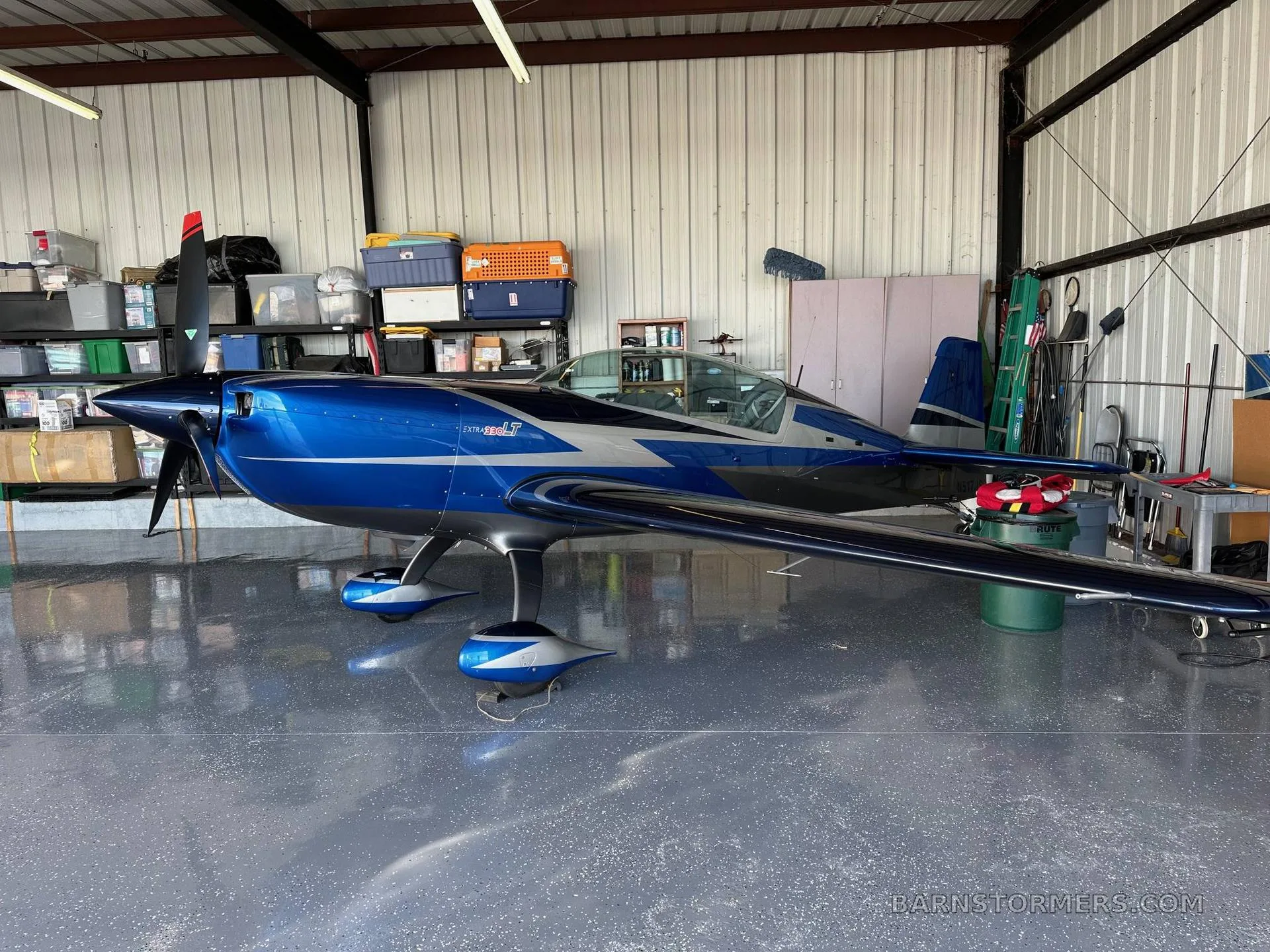 A blue and silver Extra 300LT aerobatic aircraft is parked inside a hangar with various items stored in the background.