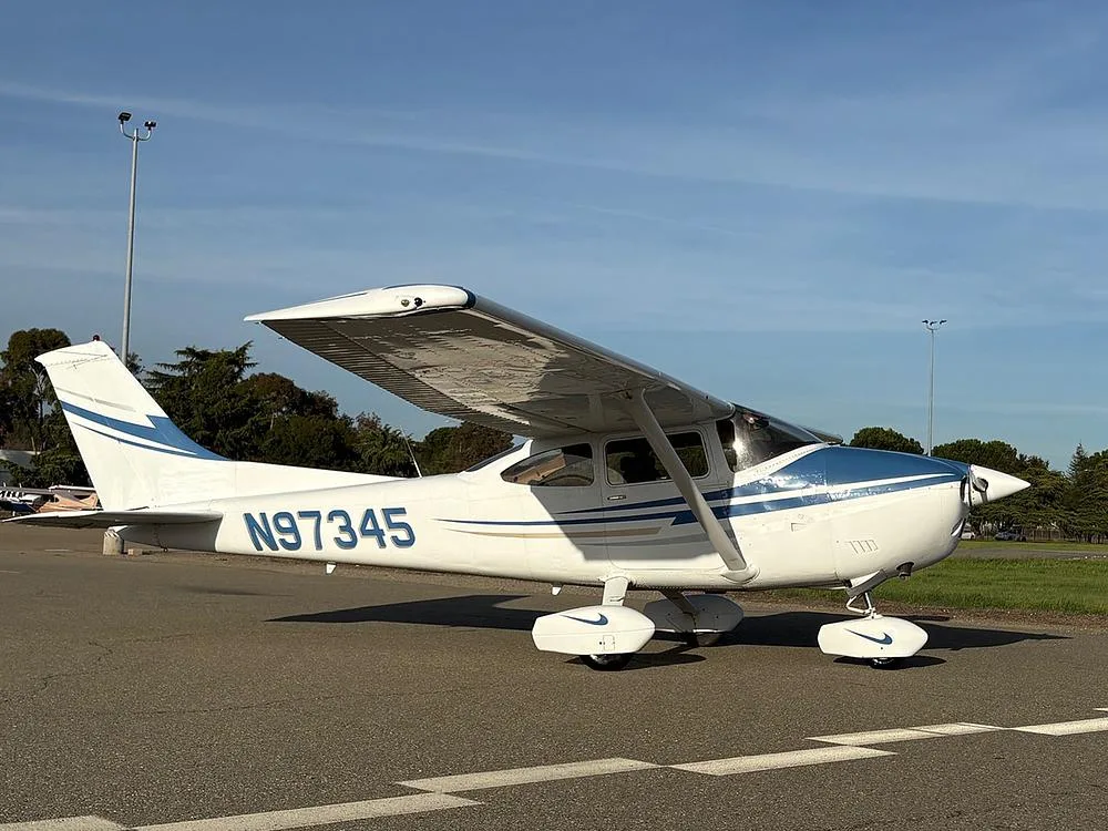 A white and blue Cessna aircraft with registration N97345 parked on an tarmac with ground markings and trees in the background.