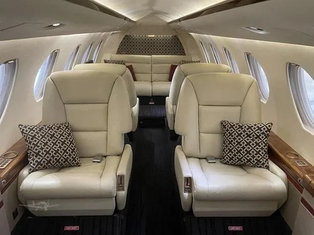 The aircraft cabin features cream leather seats with patterned pillows and dark flooring.