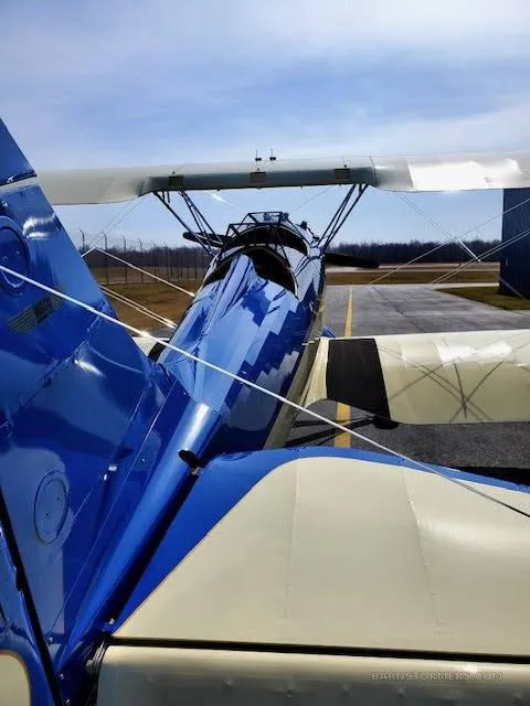 A rear view of a blue and white biplane parked on an airfield, showing its tail and upper wing.