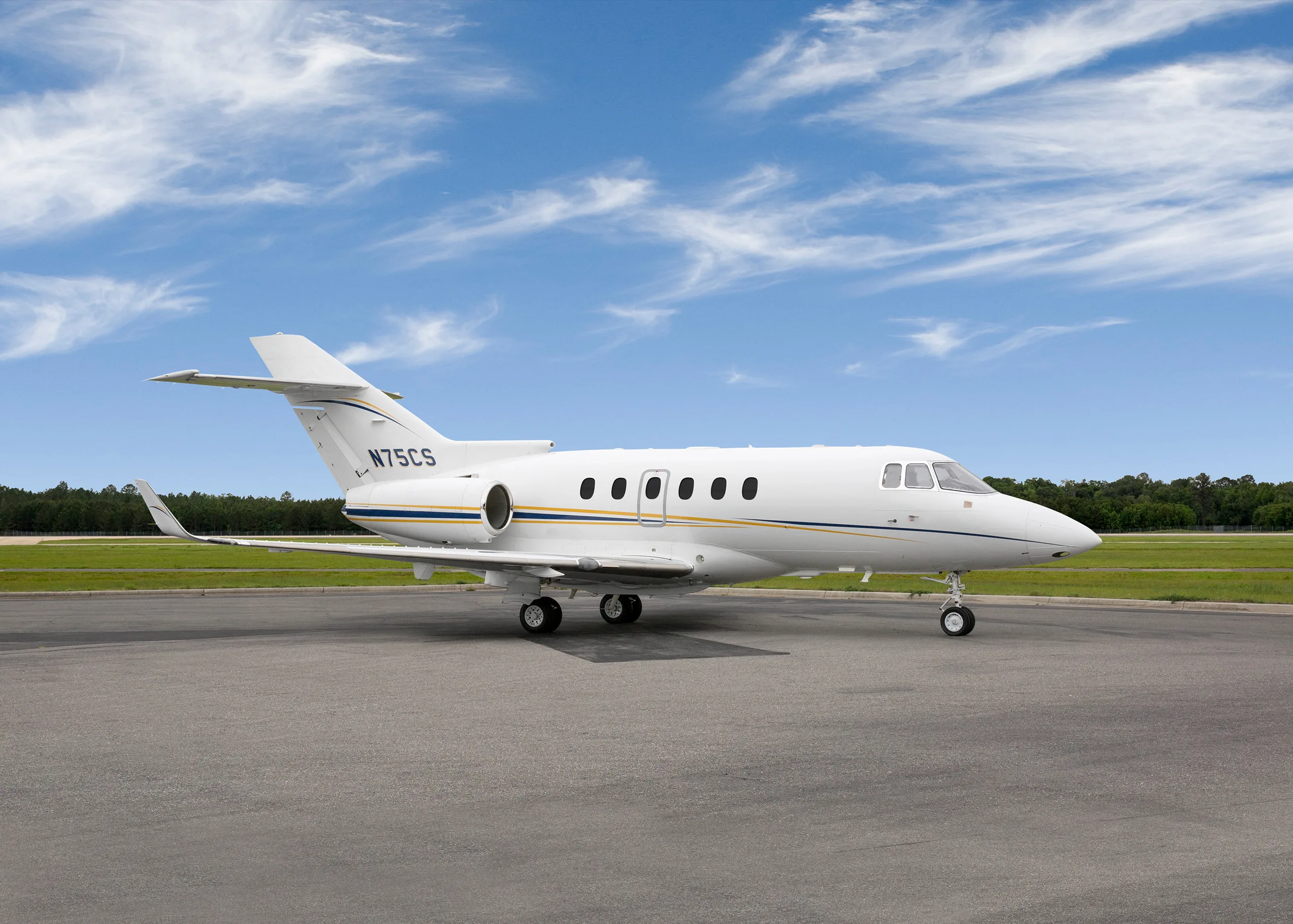 A white private jet with blue and gold stripes, registration N75CS, parked on an asphalt tarmac under a partly cloudy sky.
