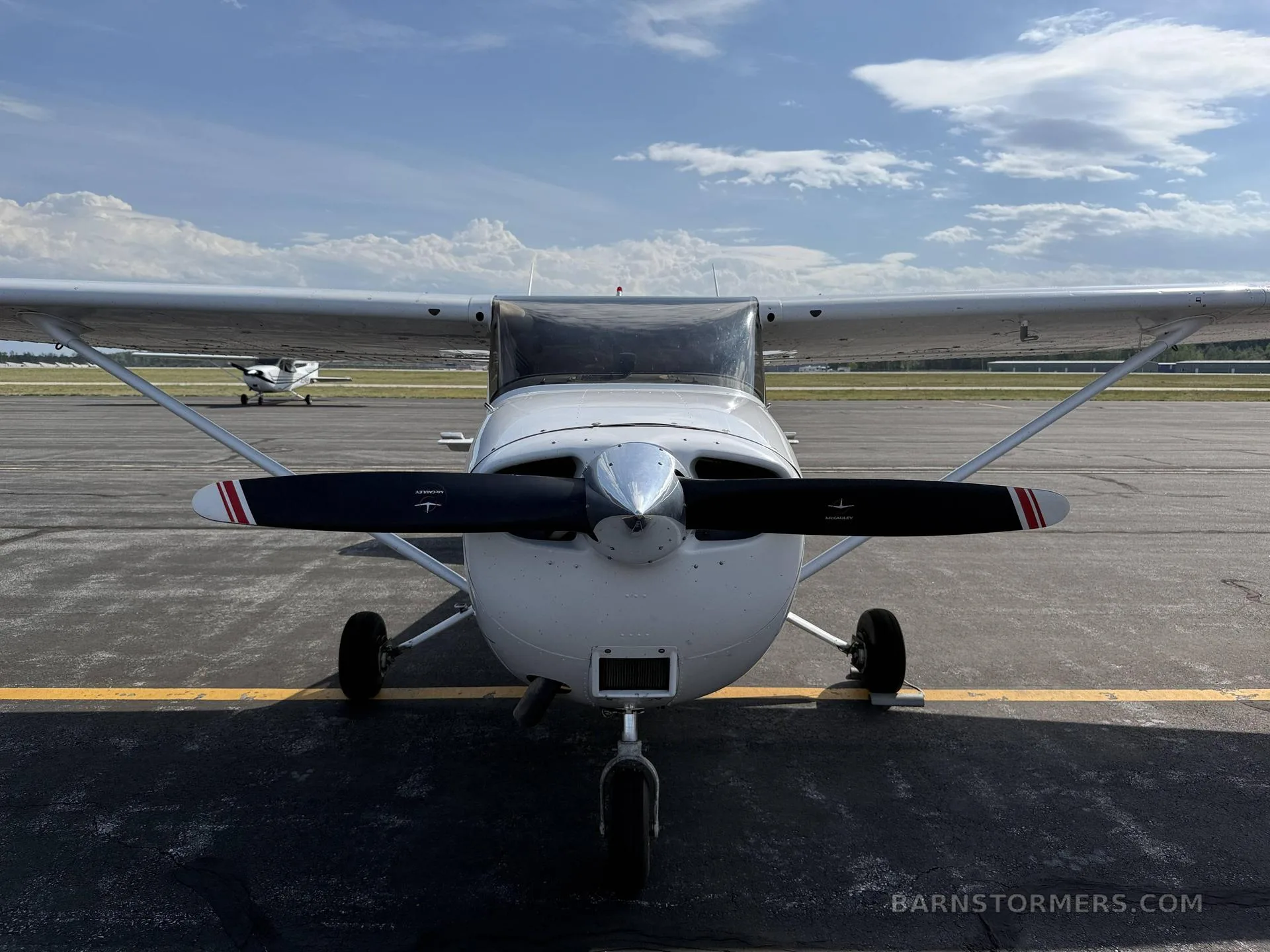 A head-on view of a white Cessna 172 aircraft with a black propeller, parked on an asphalt tarmac under a blue sky.
