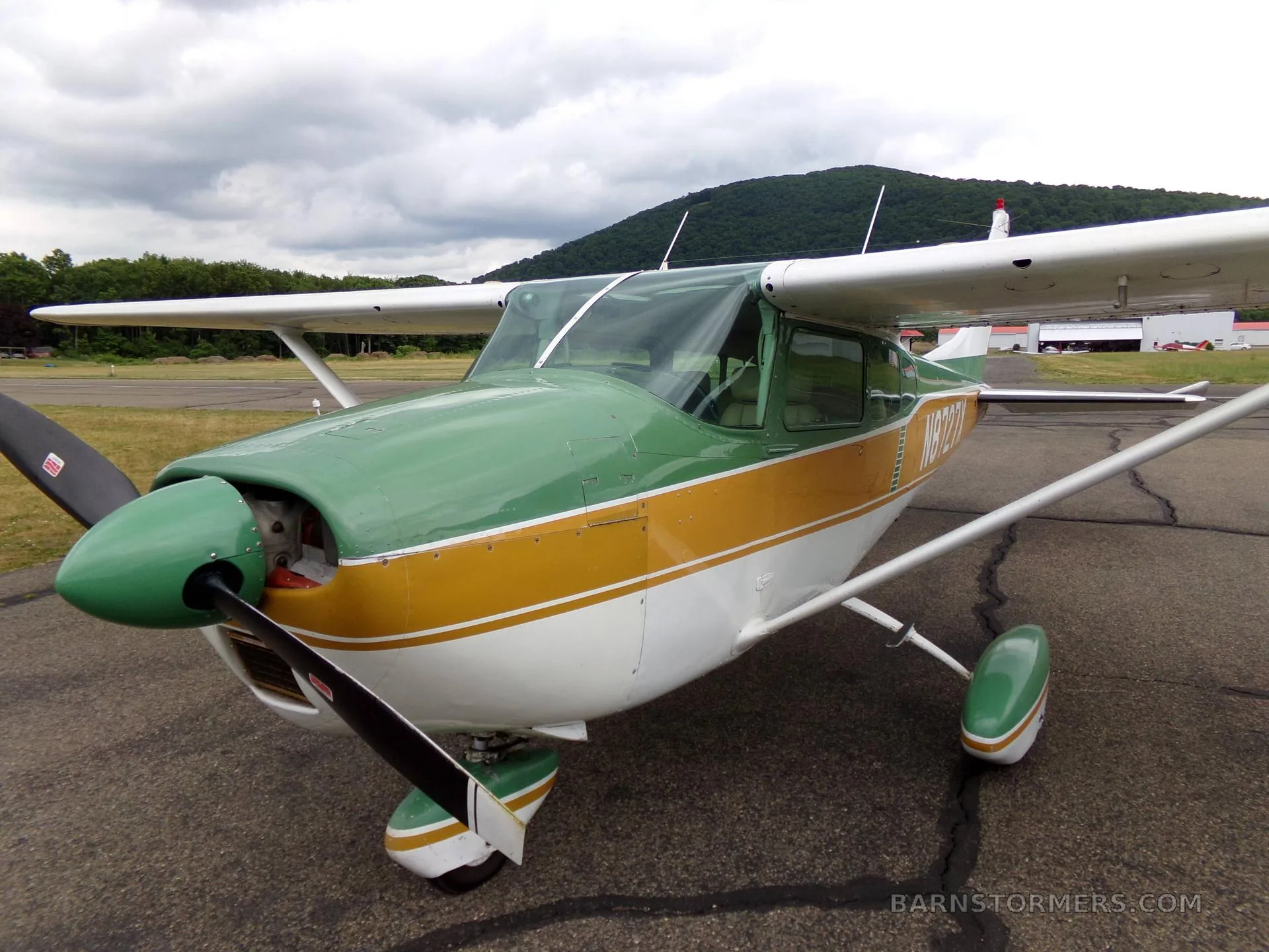 A green, gold, and white Cessna 172 aircraft with registration N8727X, viewed from the front-left on an asphalt ramp under a cloudy sky.