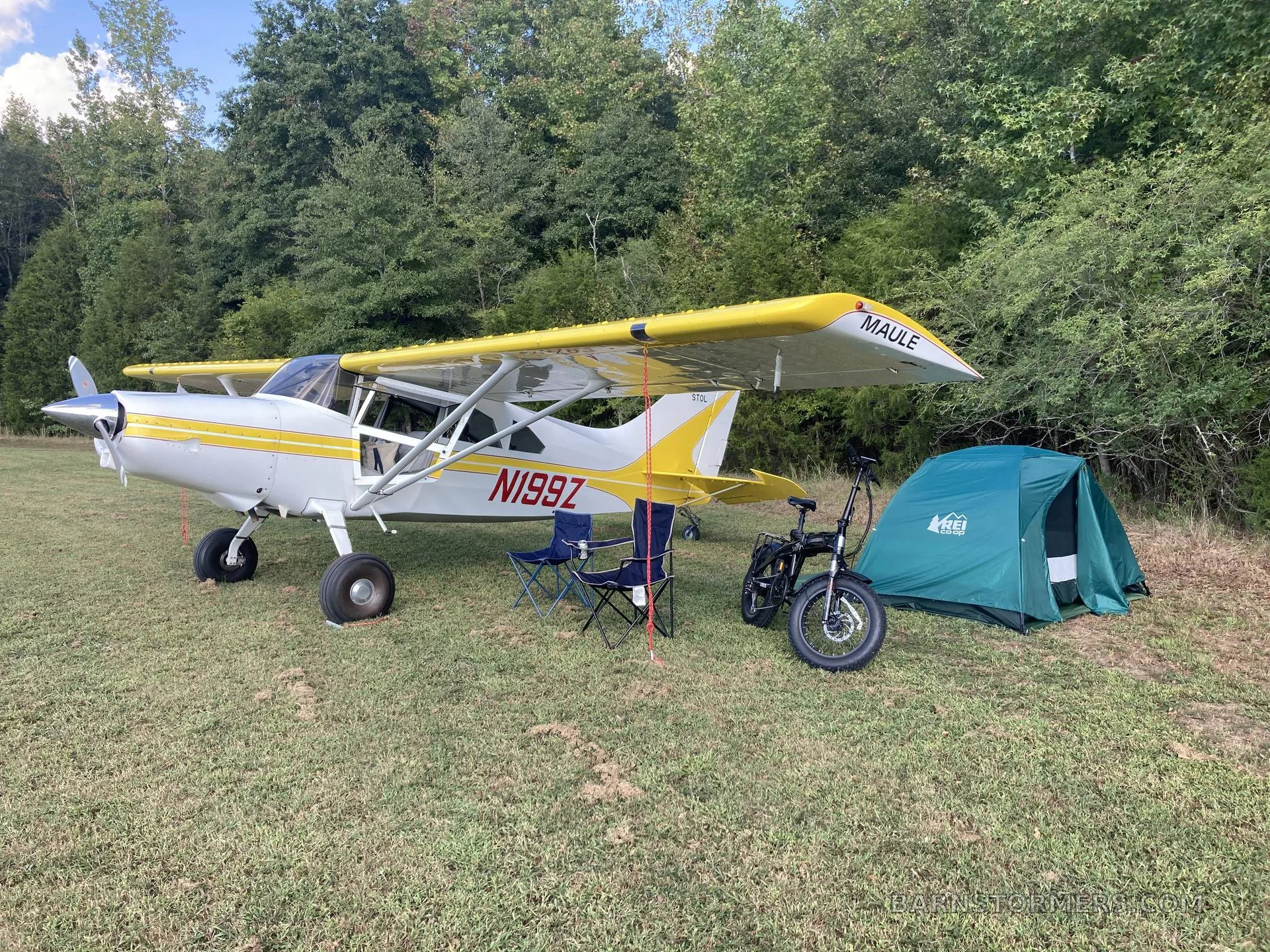 A white and yellow Maule aircraft (N199Z) is parked on a grassy field next to a green tent and an electric bicycle.