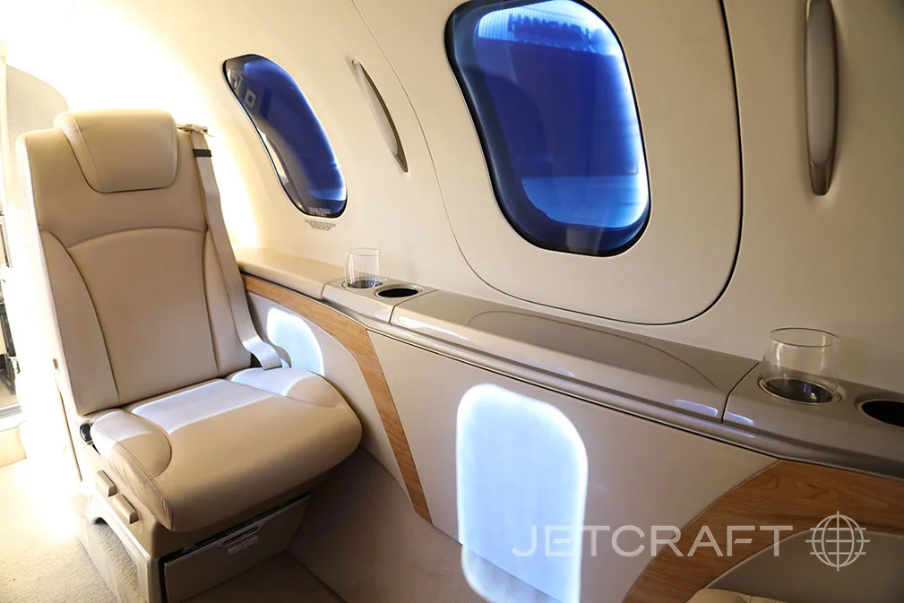 A beige leather seat and a side panel with integrated lighting and cup holders are visible inside the private jet cabin.