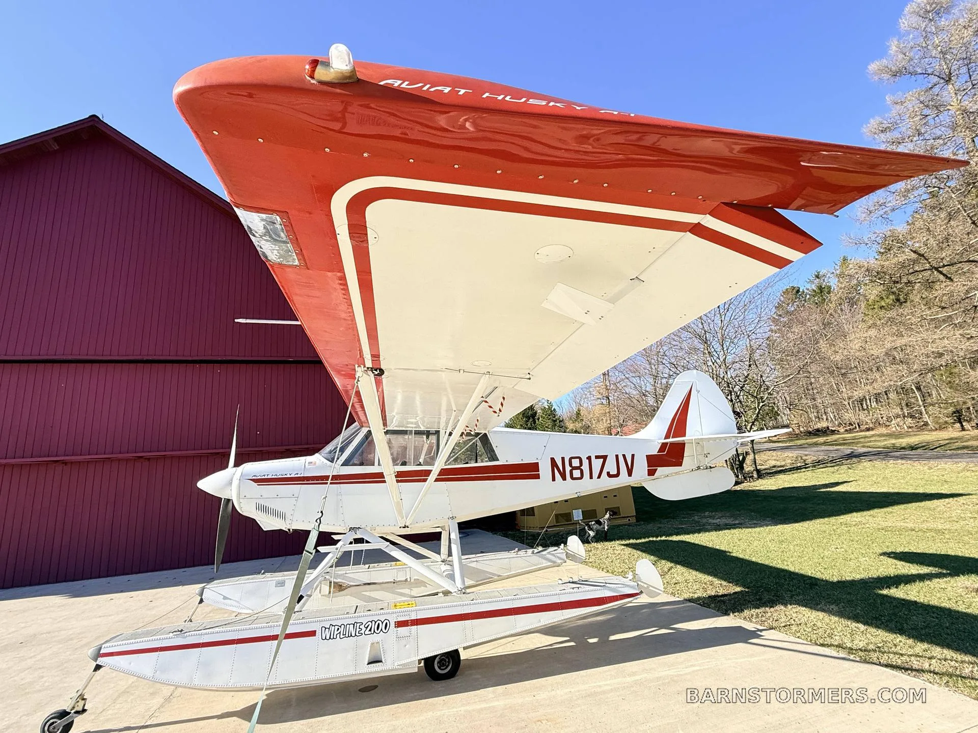 A white and red Aviat Husky A-1 floatplane (N817JV) seen from a low angle, showing the top of its red wing.